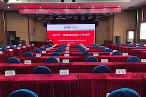 Un salón de conferencias con filas de mesas y sillas, una pantalla grande mostrando texto, y un escenario con un atril.