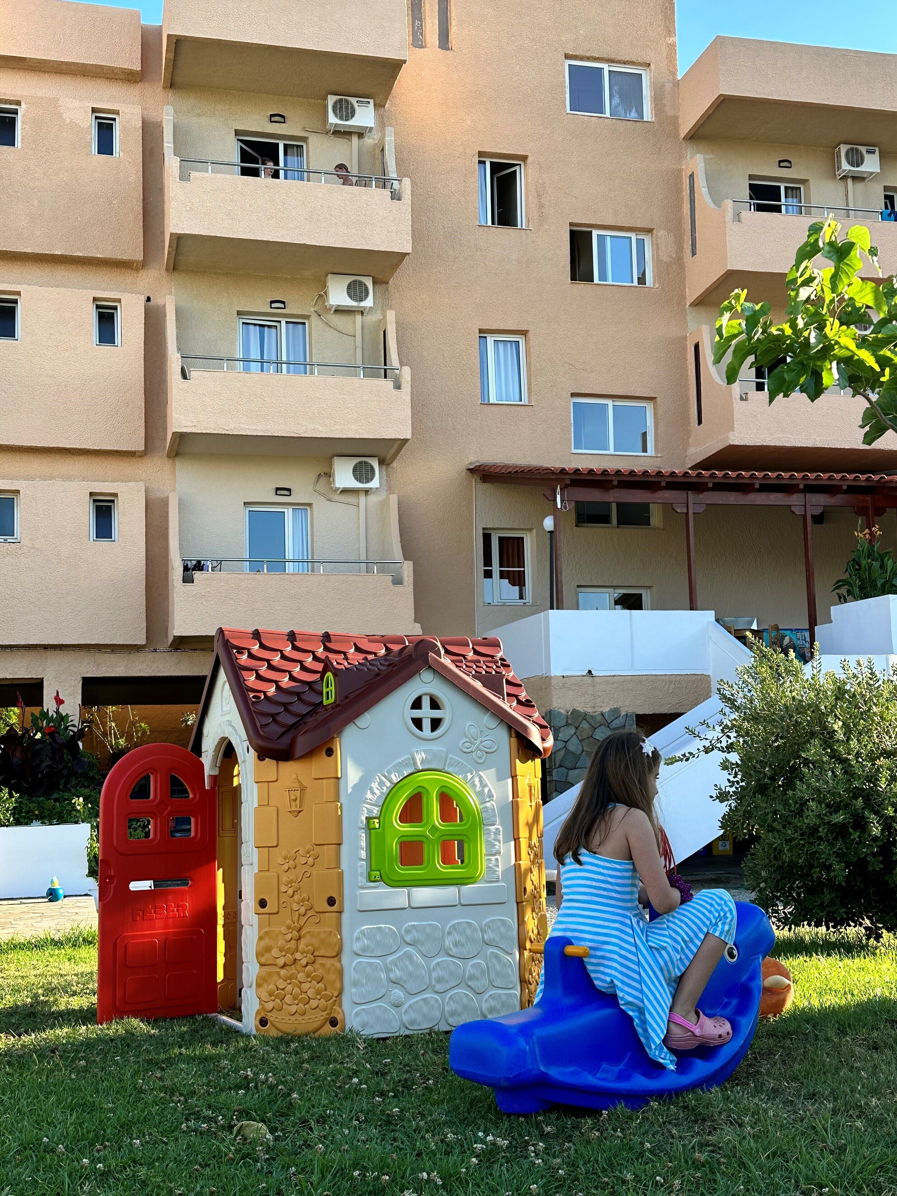 Niño en un tobogán azul frente a una casita de juegos con puerta roja y ventana verde.