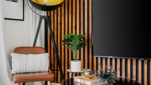 A modern living room with a brown leather chair, a white cushion, a potted plant, a glass table with decorative items, and a large black screen. - Cozy Deck • Fire Pit • EV Charger • Outdoor Dining (Austin)