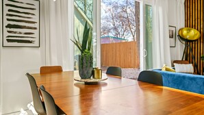 A modern living room with a wooden dining table, a blue sofa, and a large window with curtains. - Cozy Deck • Fire Pit • EV Charger • Outdoor Dining (Austin)