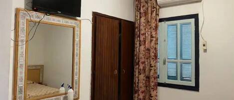 A bedroom with a television on a stand, a mirror with a decorative frame, a wardrobe, a window with shutters, and an air conditioning unit.