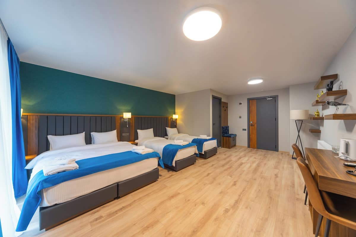 A hotel room with three beds, a wooden floor, a desk with a chair, and a small table with a lamp.