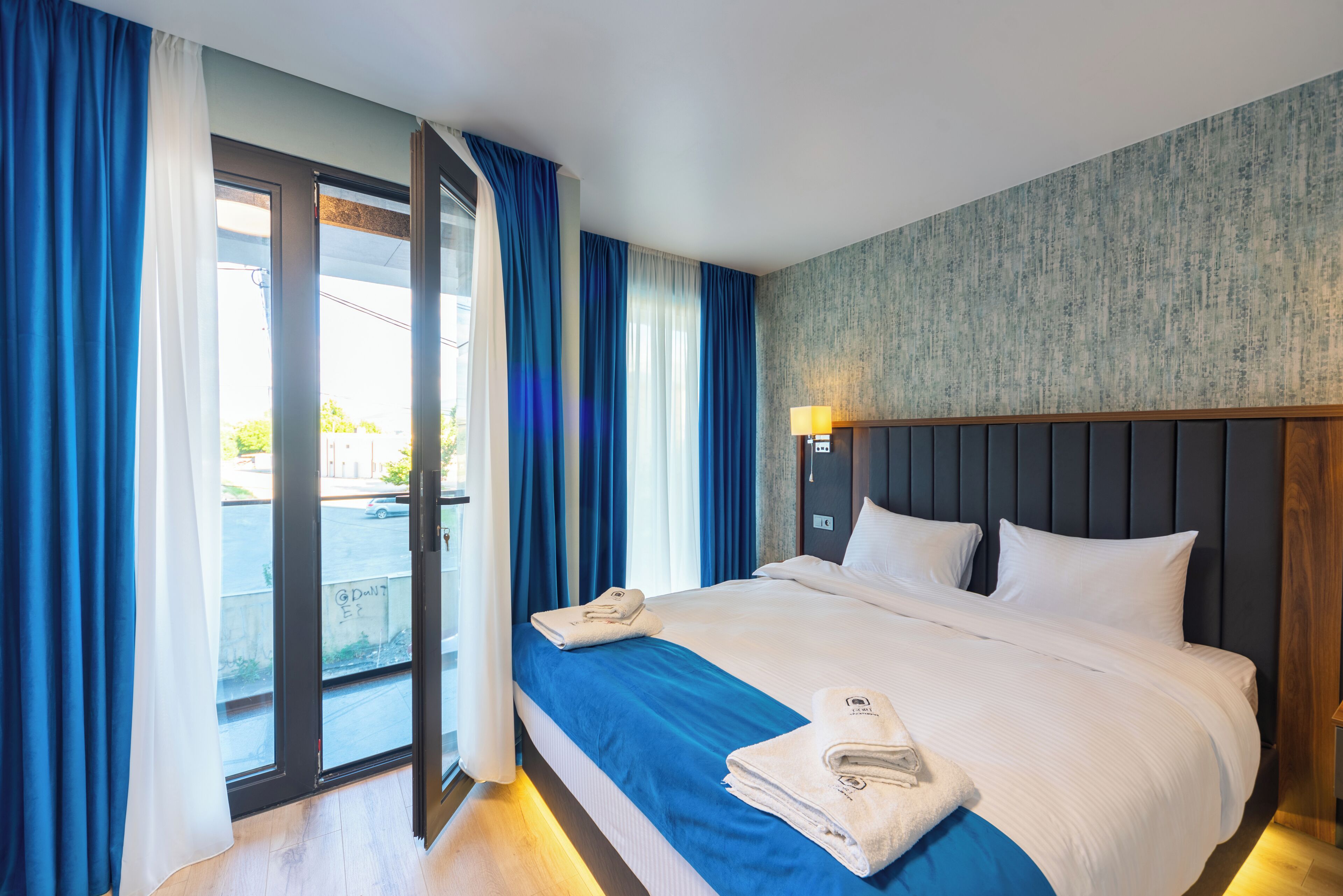 Habitación de hotel con una cama grande, vistas a través de una puerta corredera y una mesita de noche con lámpara.