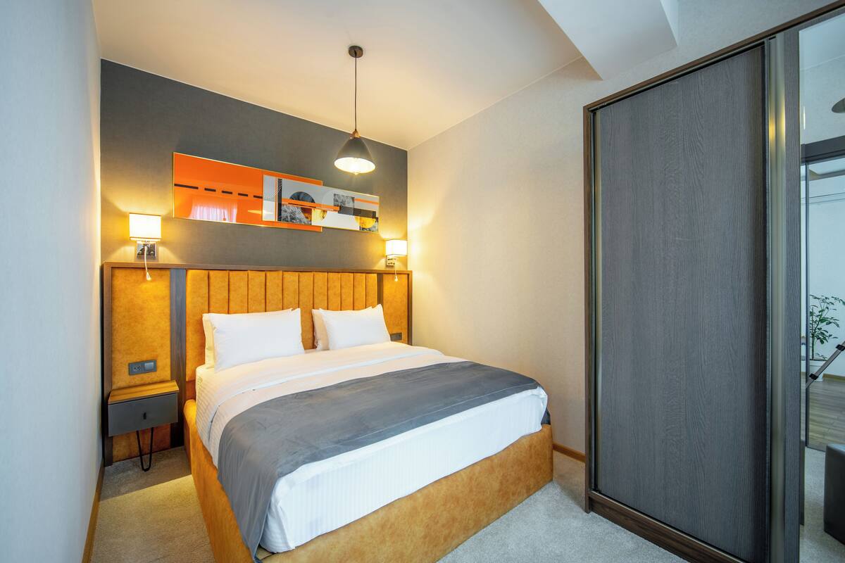 A modern hotel room with a large bed, wooden headboard, bedside lamps, a wall-mounted shelf with artwork, and a sliding glass door to the right.