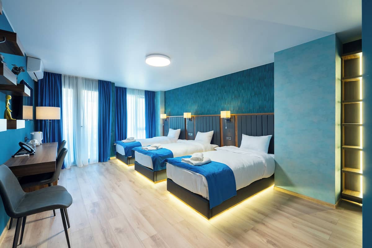 A hotel room with three beds, a desk with a chair, and a large window with curtains.