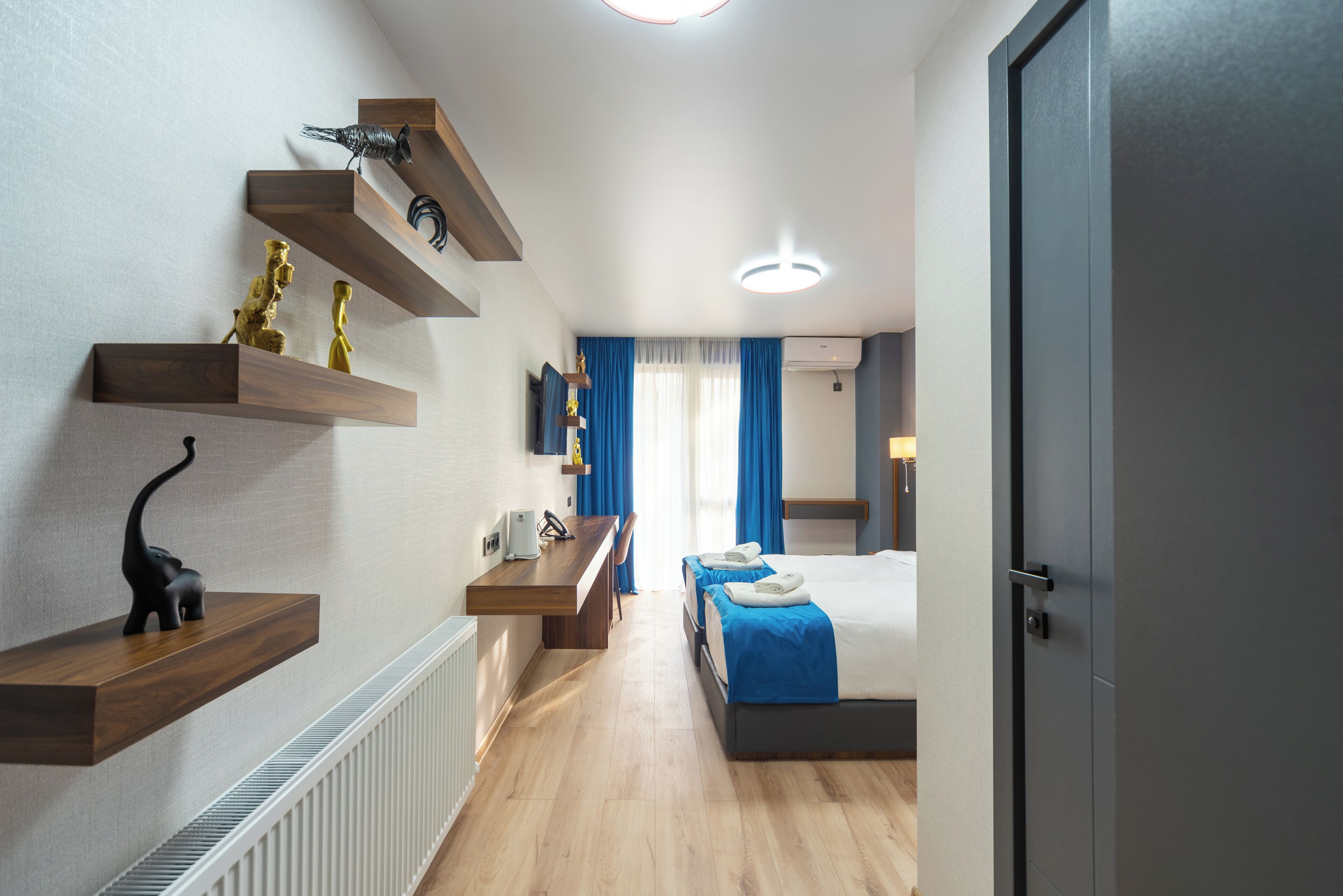 Een moderne hotelkamer met een houten bureau, een bed met blauw beddengoed en uitzicht op de gang.
