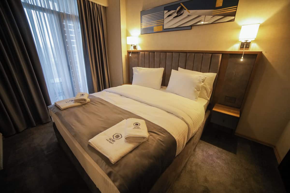 A hotel room with a large bed, two bedside tables, a headboard, and a window with curtains.