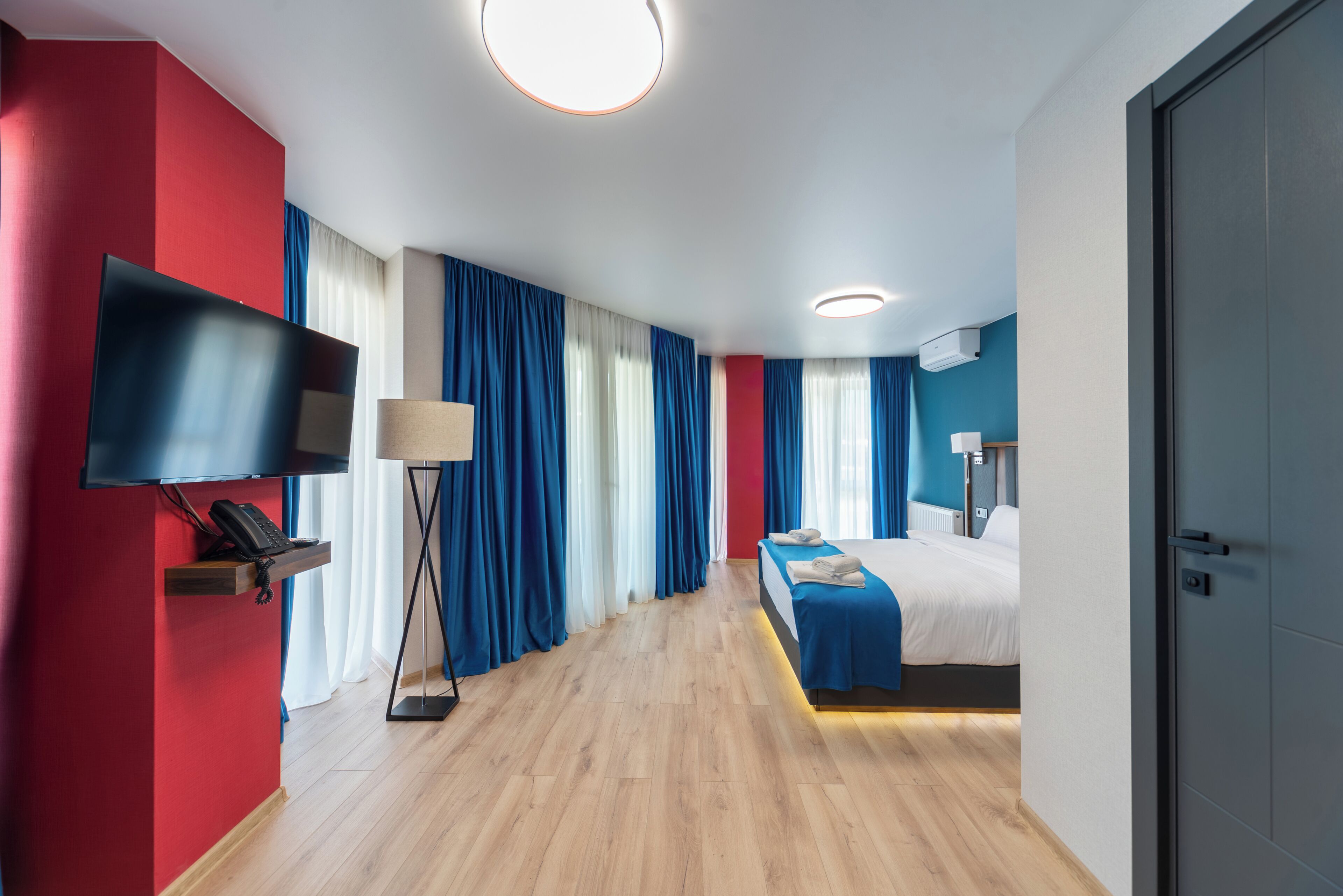 A modern hotel room with a wooden floor, a bed with a blue bedspread, a flat-screen TV mounted on the wall, and a bedside table with a lamp.