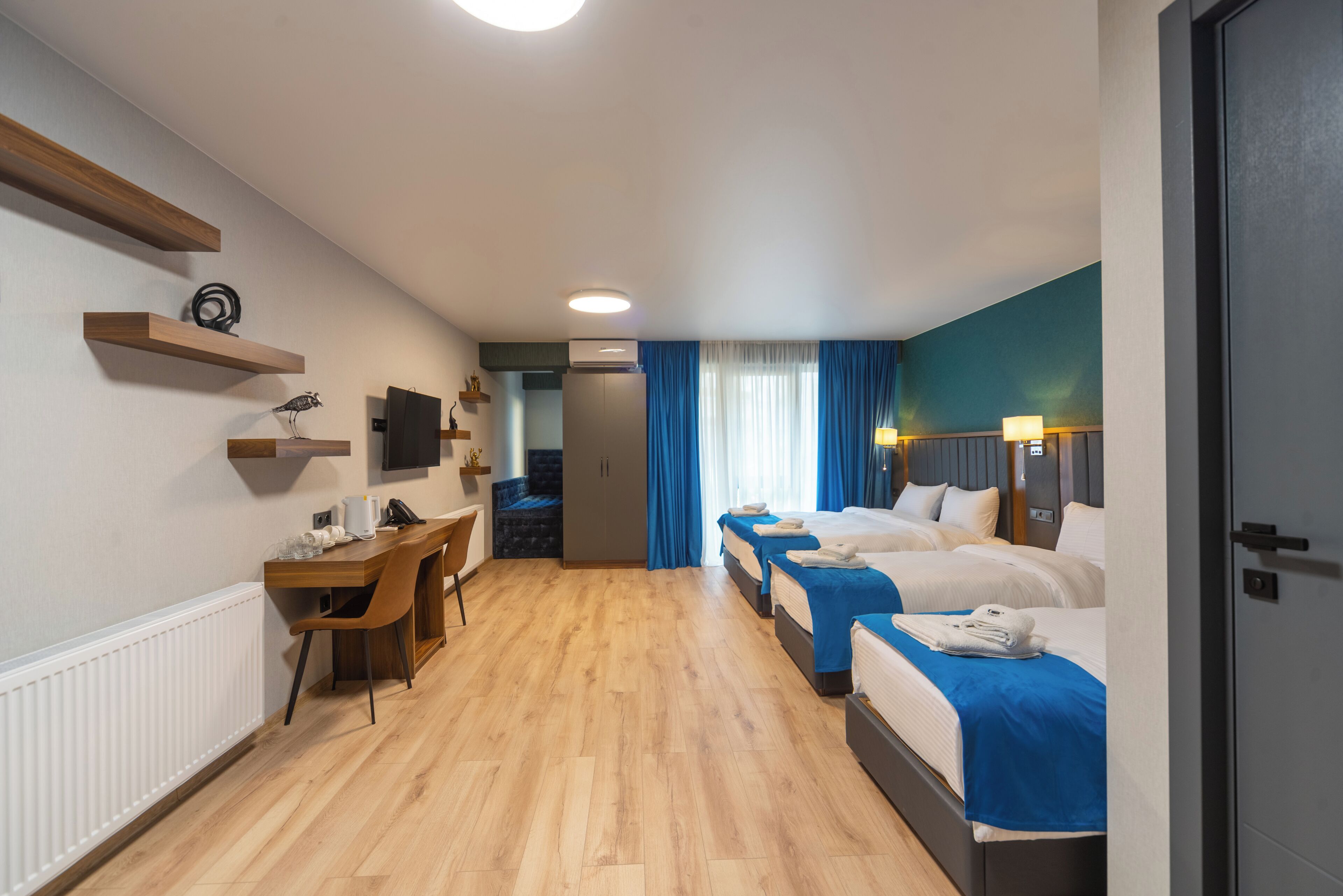 Habitación de hotel con piso de madera, un escritorio con silla, televisor montado en la pared y tres camas con ropa de cama azul.