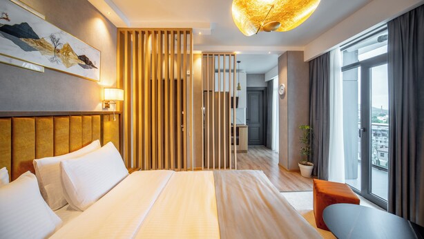 Una habitación de hotel moderna con una cama grande, un escritorio, una silla, una planta en maceta y vistas a la ciudad desde la ventana.