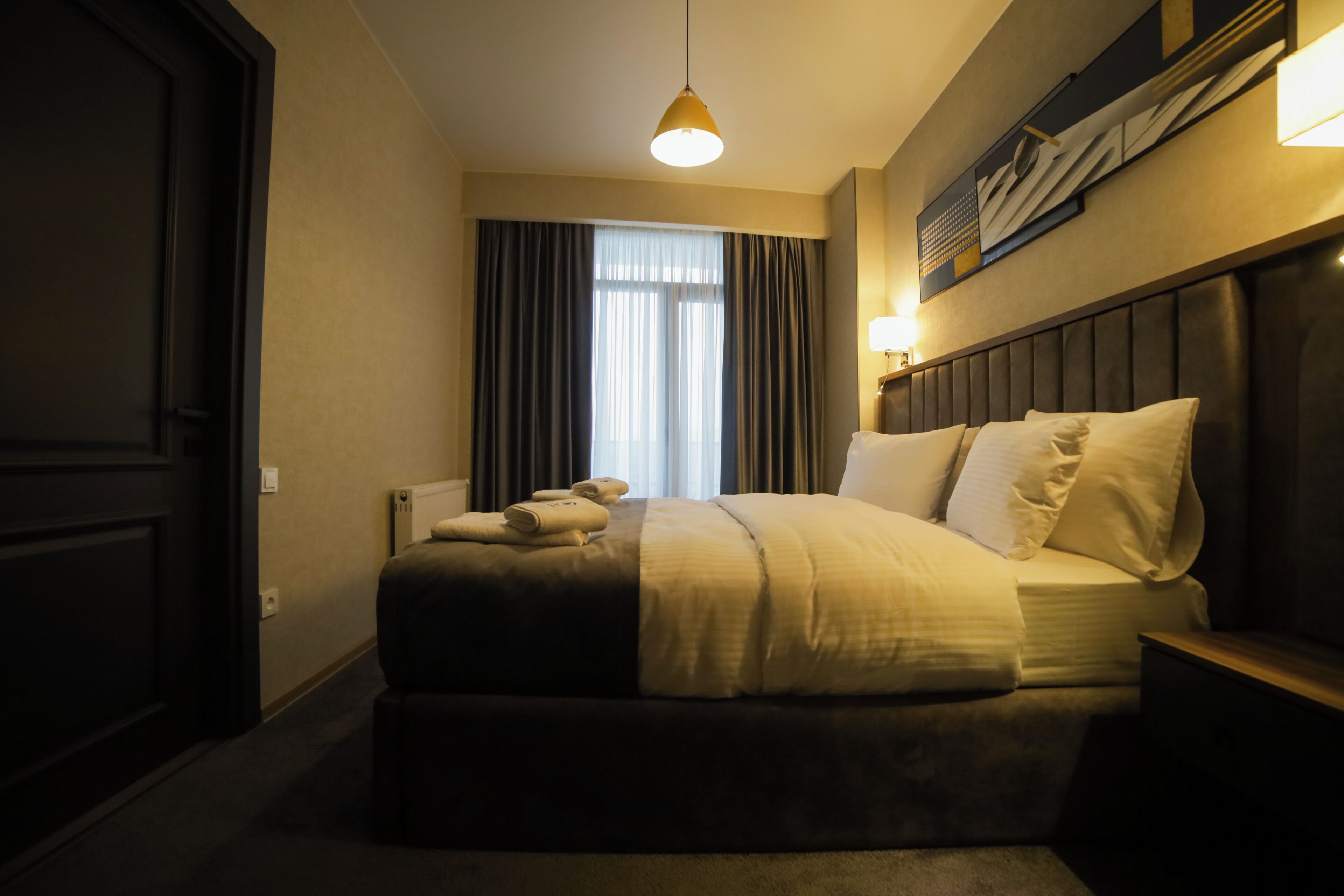 A hotel room with a large bed, a headboard, a bedside table, a lamp, and a window with curtains.