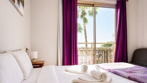Ein Hotelzimmer mit einem Bett, einem Nachttisch, einer Lampe und Blick auf Palmen durch das Fenster.