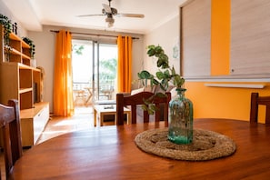 Un espace repas comprenant une table en bois, des chaises, un ventilateur de plafond et une vue sur l’extérieur, accessible par une porte vitrée.