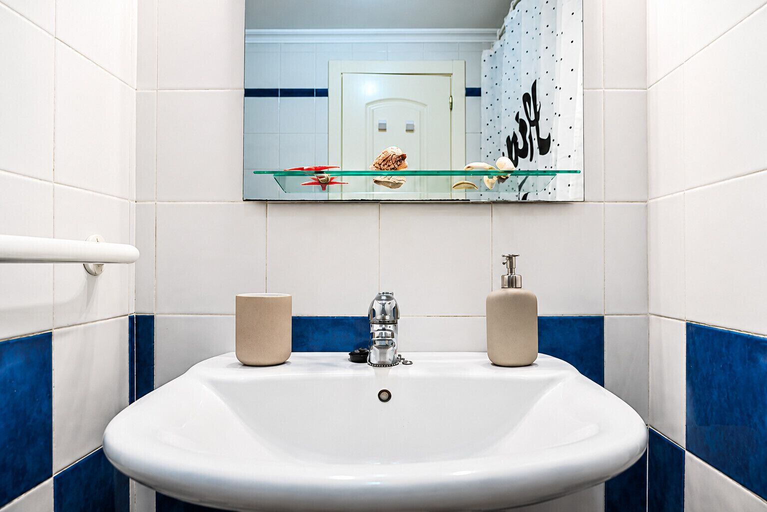 Een badkamer met een witte wastafel, twee zeepdispensers, een glazen plankje met decoratieve voorwerpen, en een spiegel die een deur en een behang met patroon weerspiegelt.