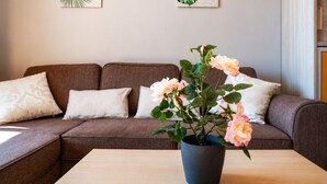 Ein Wohnzimmer mit einem braunen Sofa, zwei weißen Kissen, einem Holztisch und einem Blumentopf mit rosa Blüten.