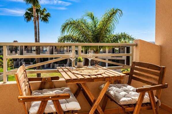 A wooden table and chairs on a balcony with a view of palm trees and a building.