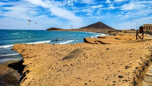 Ein Strand mit felsiger Küste, einem hölzernen Steg und einem Berg im Hintergrund.