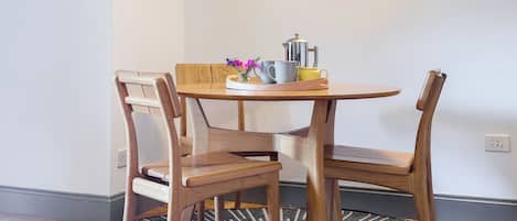 A dining area with a round wooden table, two wooden chairs, a coffee pot, and a vase with flowers.