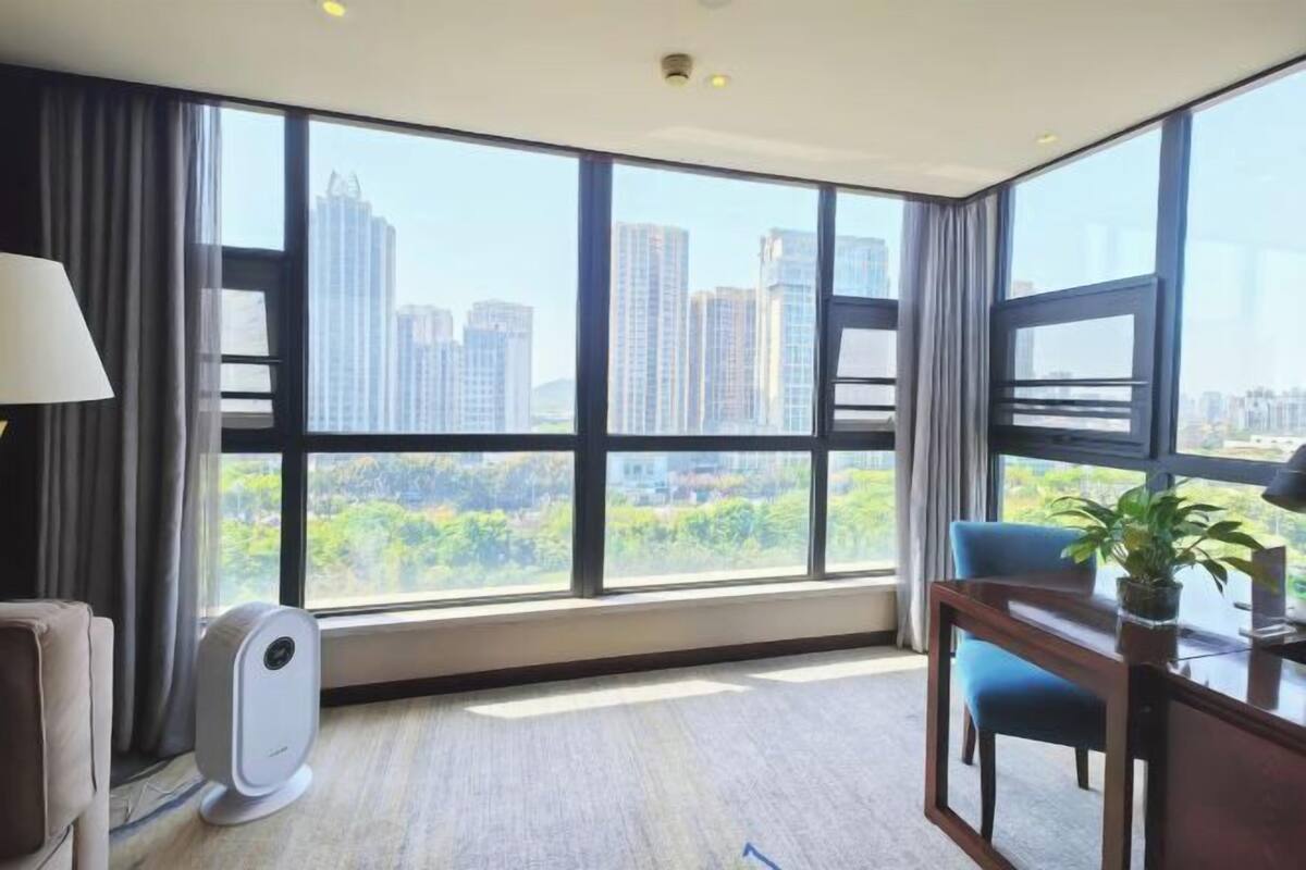 A hotel room with large windows, a standing heater, a blue chair, and a desk with a plant.