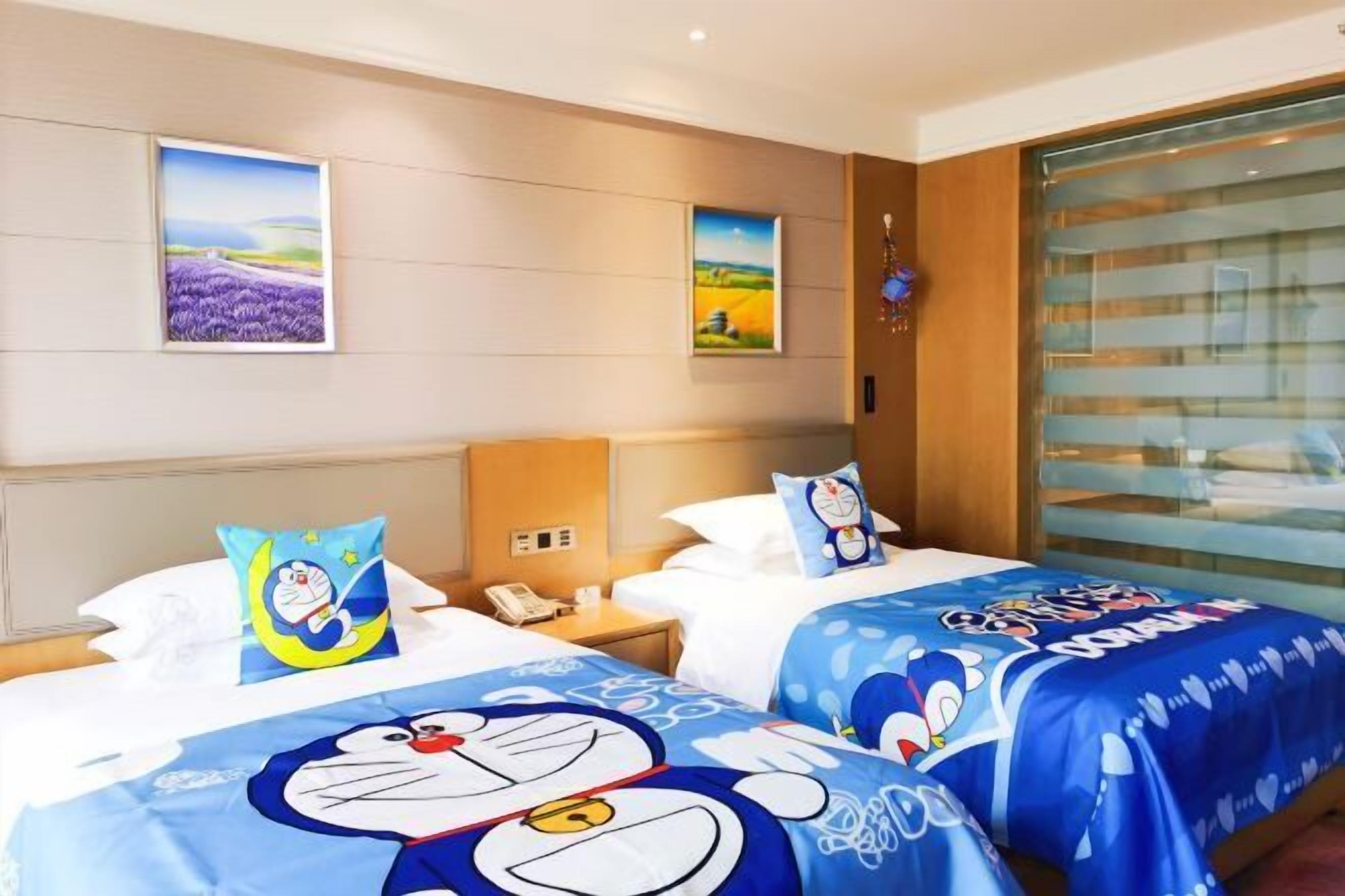 A hotel room with two beds, each with a blue blanket featuring a cartoon character, a nightstand with a phone, and wall art depicting landscapes.