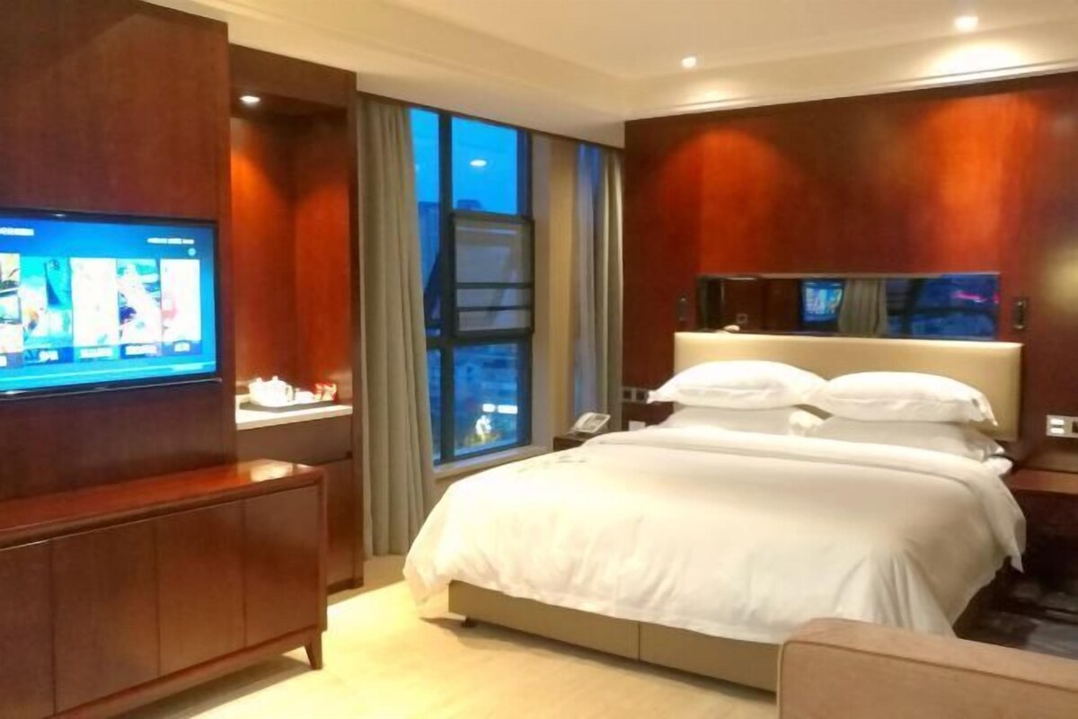 A hotel room with a large bed, a TV on the wall, and a window with curtains.
