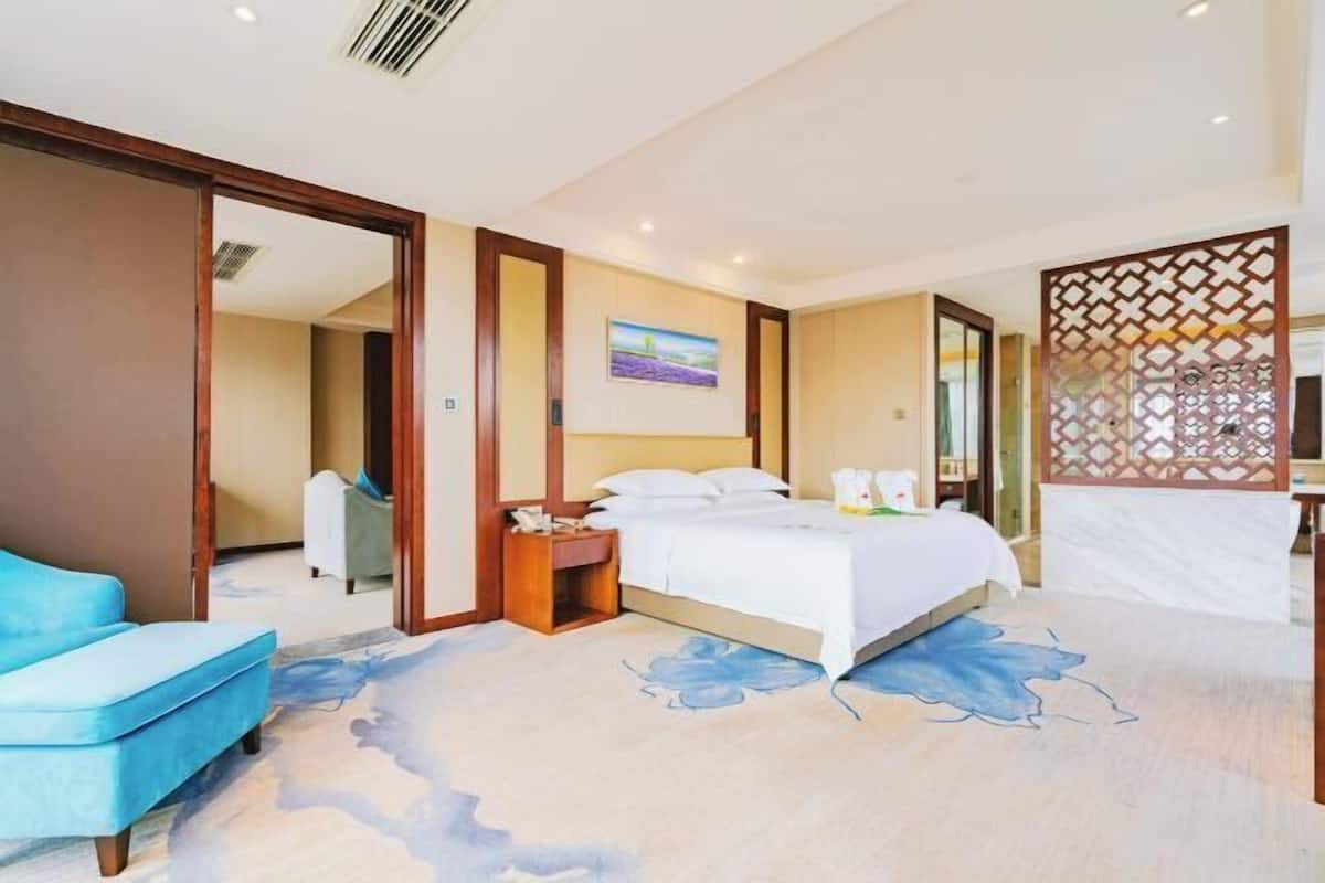 A hotel room with a bed, a blue chair, a nightstand, a painting on the wall, and a decorative screen.