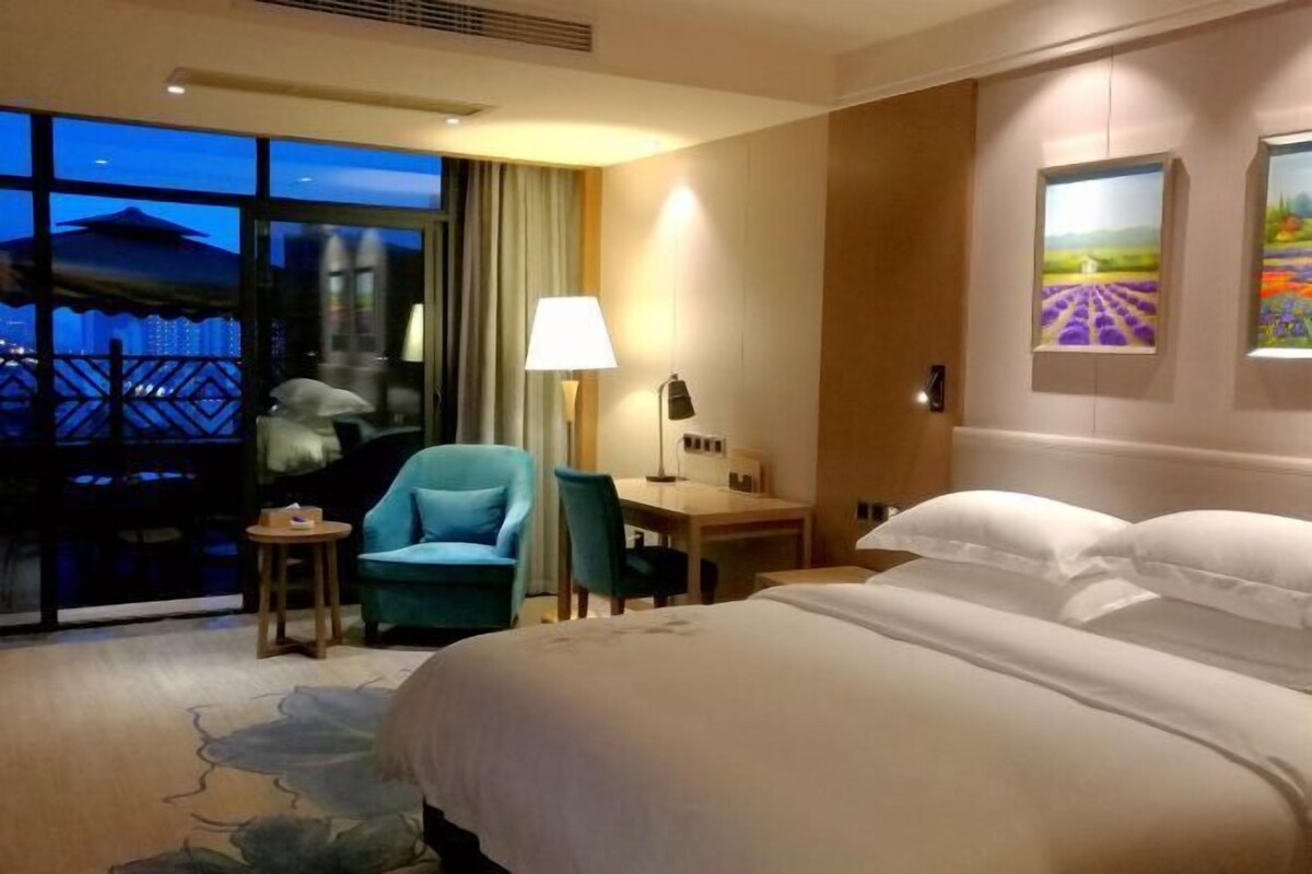 A hotel room with a large bed, a desk, a chair, and a view of a cityscape through large windows.