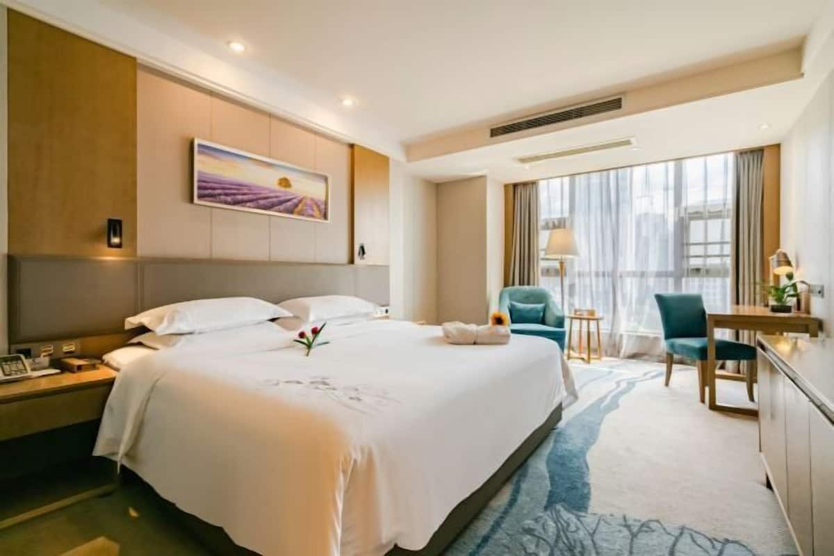 A hotel room with a large bed, a desk with a lamp, a chair, a window with curtains, and a painting on the wall.