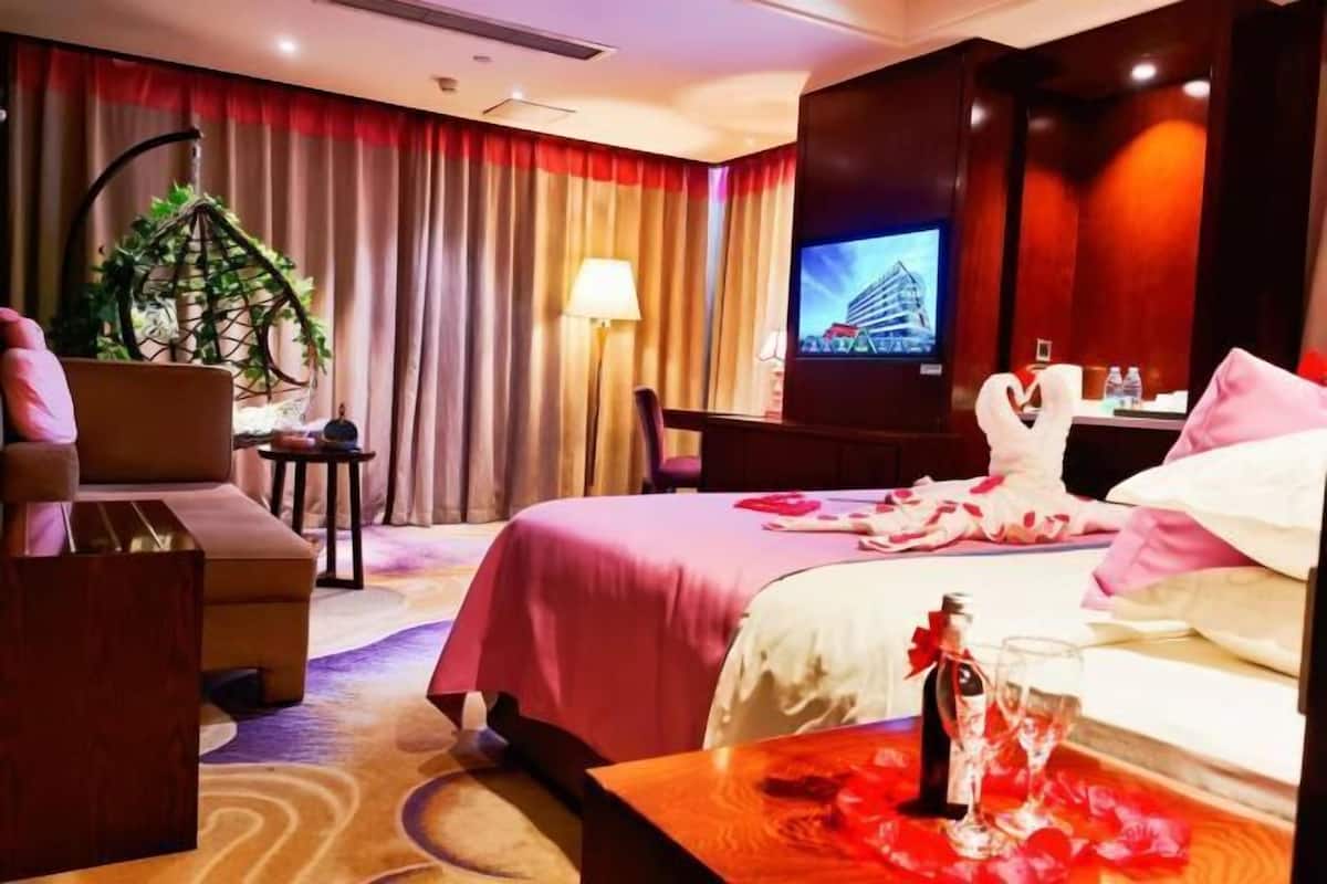 A hotel room with a bed, a sofa, a TV, a plant, and a table with a bottle and glasses.
