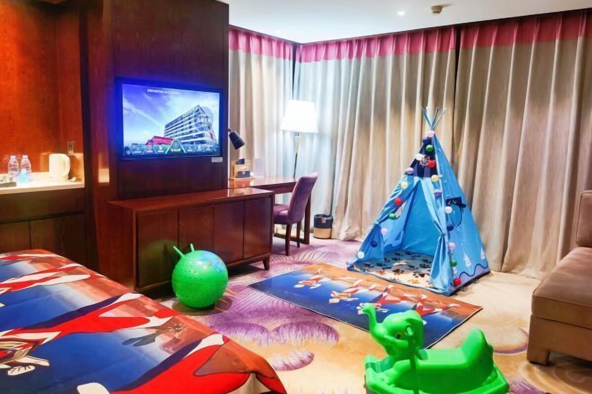 A hotel room with a TV mounted on the wall, a desk with a chair, a teepee, and children's toys on the floor.