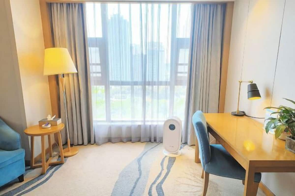 A hotel room with a bed, a bedside table with a lamp, a window with curtains, a dining table with chairs, and a small table with a plant.