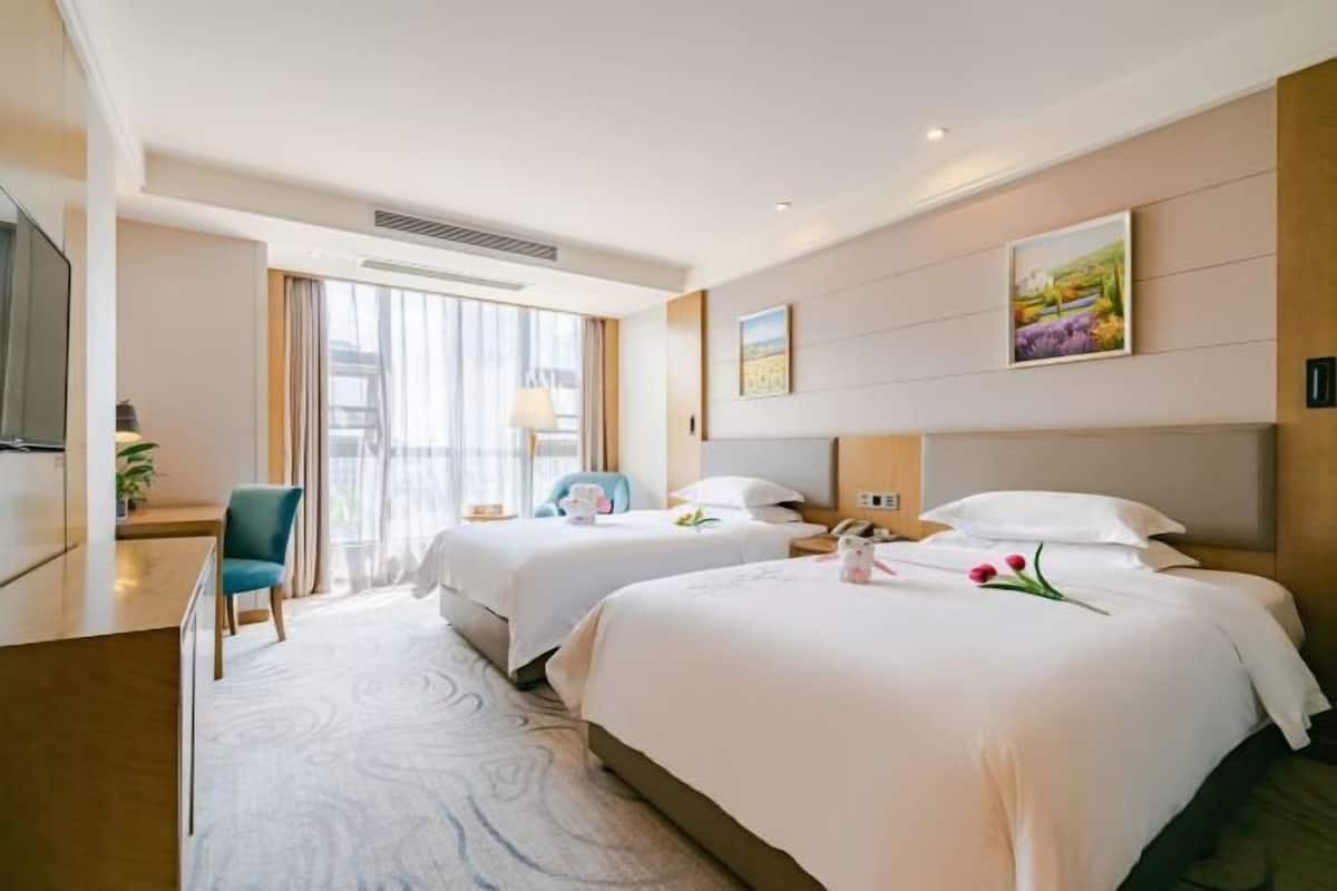 A hotel room with two beds, a desk, a chair, and a large window with curtains.