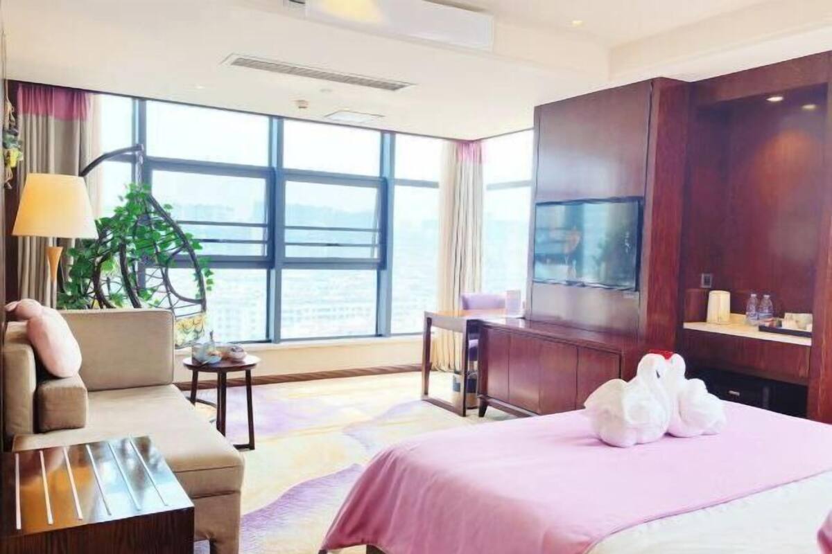 A hotel room with a bed, a sofa, a desk, a TV, and a large window offering a city view.