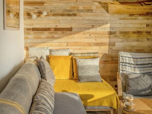 A cozy room with a wooden wall, a bed with a yellow blanket, a grey sofa, and a chair with a knitted throw.