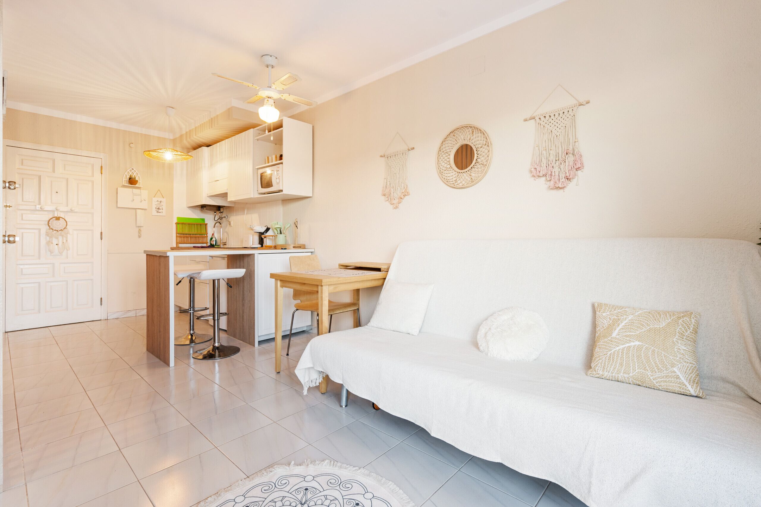 A cozy, well-lit room with a white sofa, a wooden dining table, and a kitchen area.