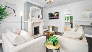 A living room with a fireplace, white sofas, a coffee table, and a TV mounted on the wall. - The unit comes high-speed WiFi, hotel-quality linen, and toiletries for your comfort. (Birmingham)