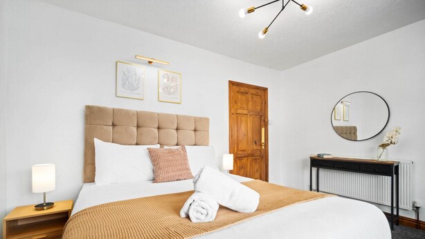 A neatly made bed with a tufted headboard, a bedside table with a lamp, a wooden door, a round mirror, and framed artwork on the wall. - The unit comes high-speed WiFi, hotel-quality linen, and toiletries for your comfort. (Birmingham)