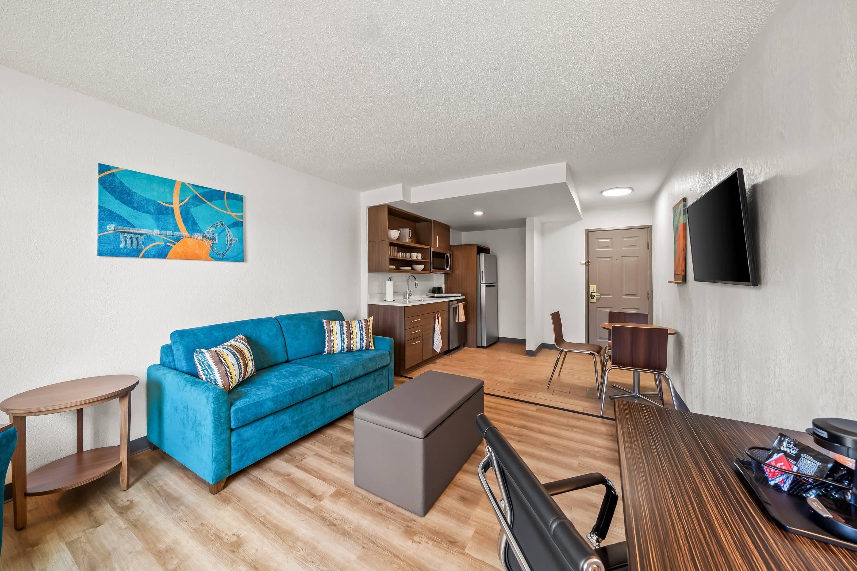 A compact living space with a sofa, ottoman, small table, kitchenette, and a flat-screen TV mounted on the wall.