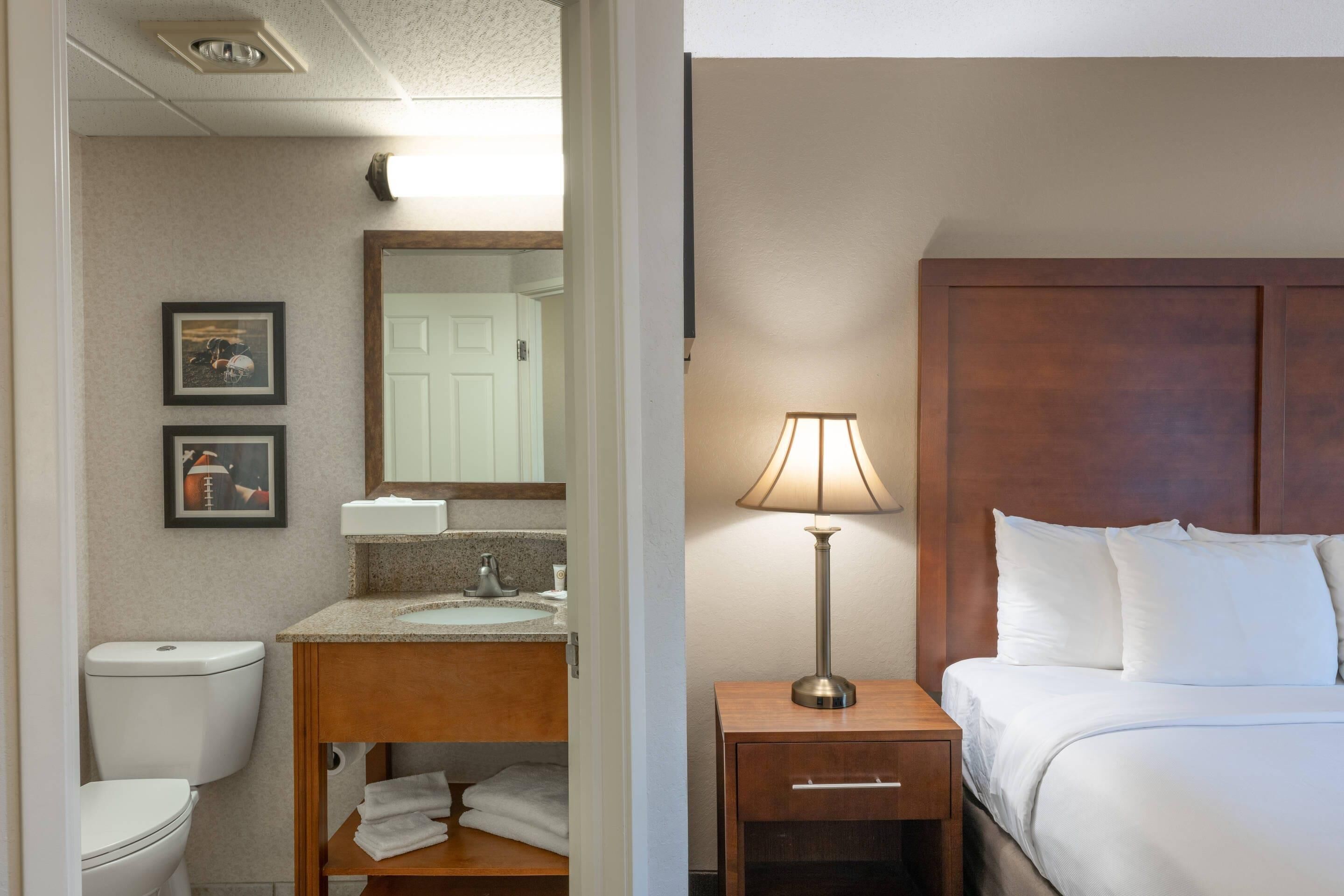A hotel room with a bed, a bedside lamp, a wooden nightstand, a bathroom with a toilet, sink, and mirror.