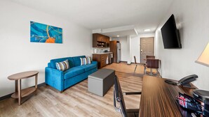 A modern living room with a blue sofa, a small table, a grey ottoman, a wooden desk with a lamp, and a kitchen in the background. - MainStay Suites Foxboro – Mansfield (Foxborough)