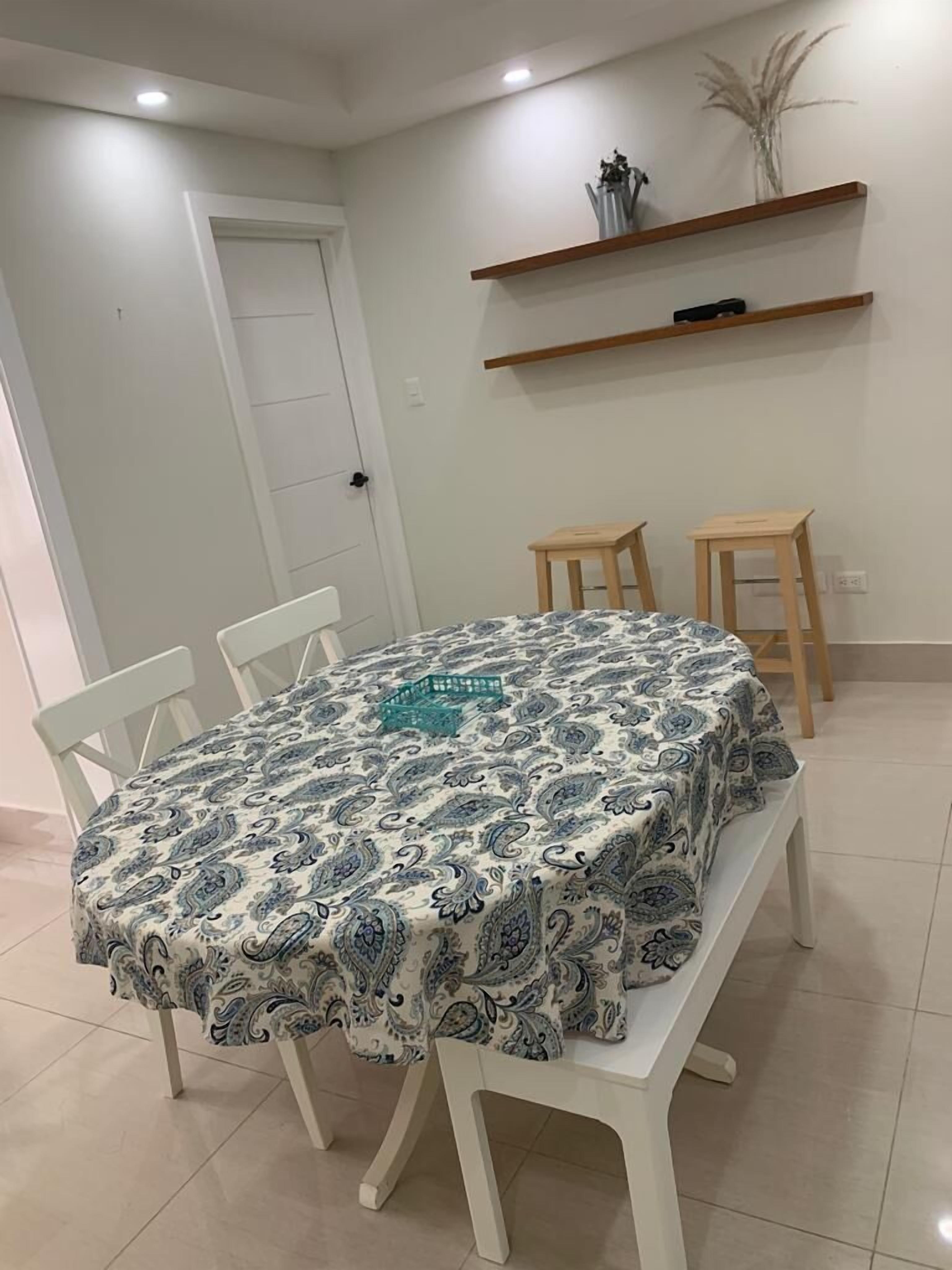 Ein Essbereich mit einem Tisch, der mit einer gemusterten Tischdecke bedeckt ist, umgeben von weißen Stühlen. Zwei hölzerne Hocker und ein kleiner Tisch mit einer Vase stehen auf einem Regal darüber.