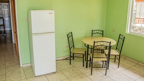 A kitchen with a white refrigerator, a dining table with four chairs, and a window with a curtain. - Charlie's Cottages (Negril)