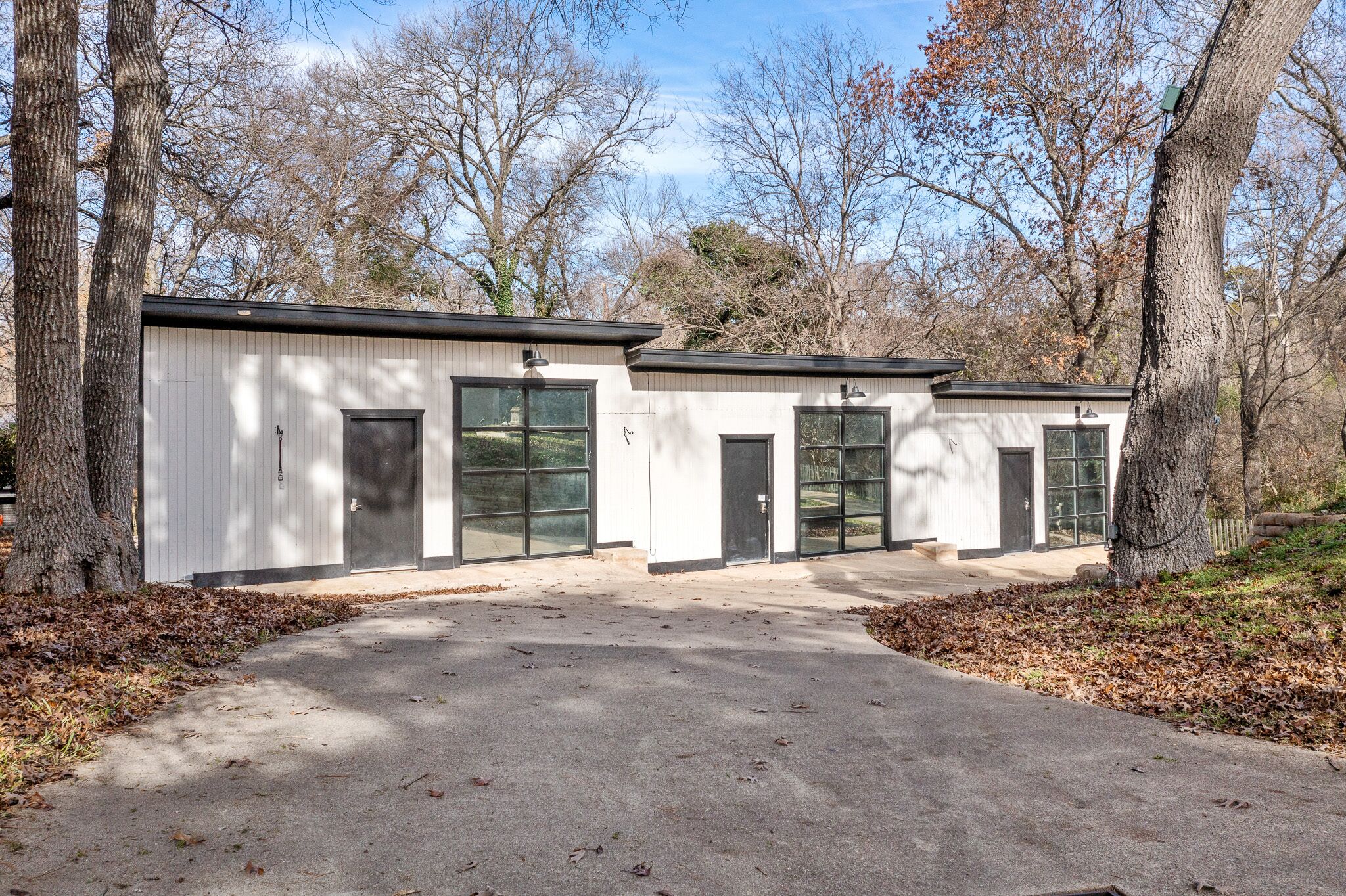 A row of three white buildings with large windows and doors, surrounded by trees and a paved area.