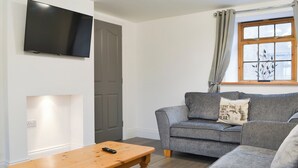 Ett vardagsrum med en grå soffa, ett soffbord i trä, en platt-TV och ett fönster med gardiner.