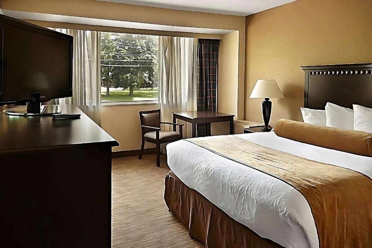 Quarto de hotel com uma cama, uma escrivaninha com televisão, uma cadeira, um abajur e uma janela com cortinas.