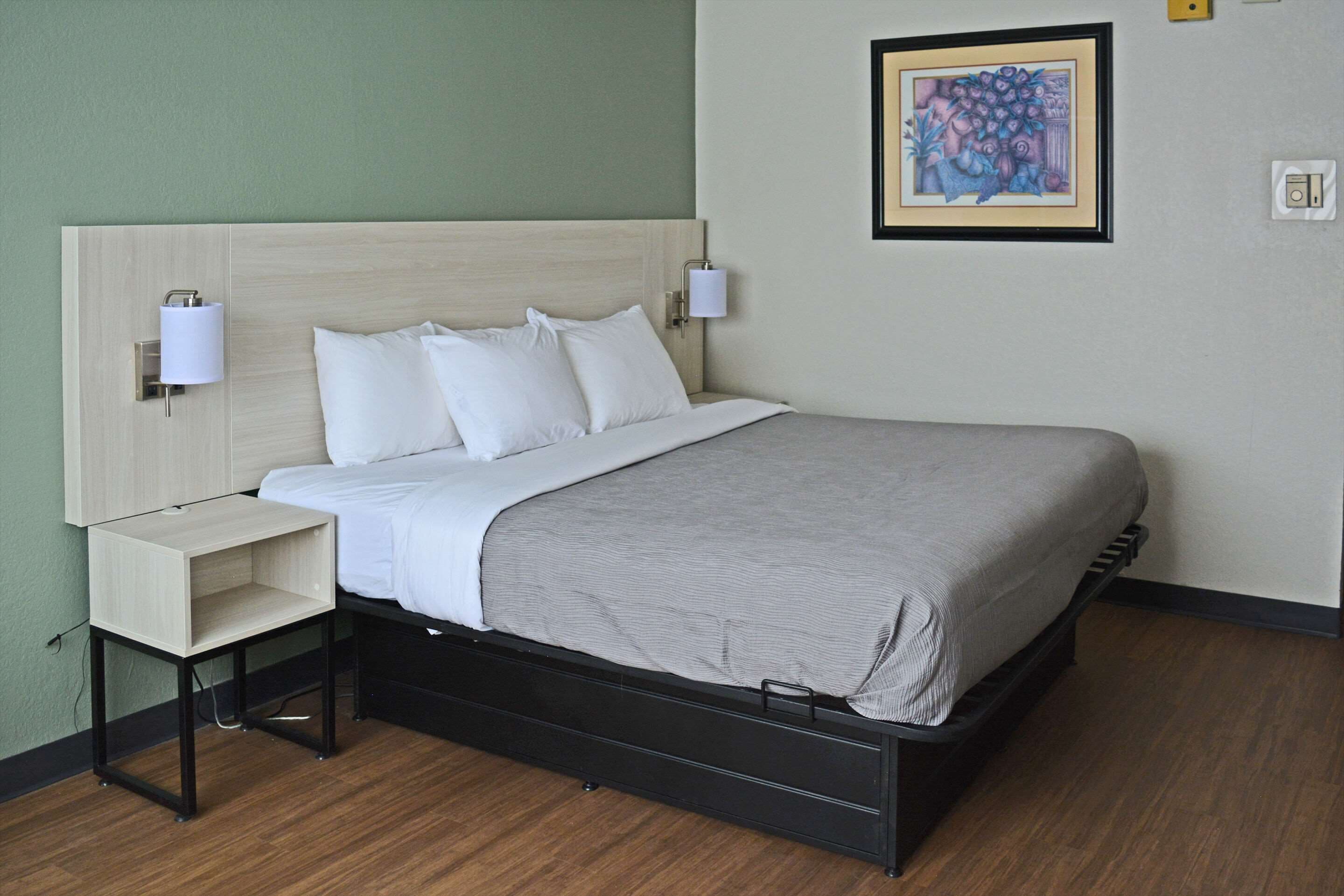 A neatly made bed with white linens and a gray blanket, a bedside table with a lamp, and a framed artwork on the wall.