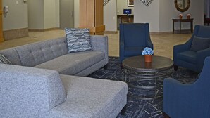 A lobby with a grey sofa, blue armchairs, a round coffee table, and a small table with a flower vase. - Quality Inn & Suites Syracuse - Carrier Circle (East Syracuse)