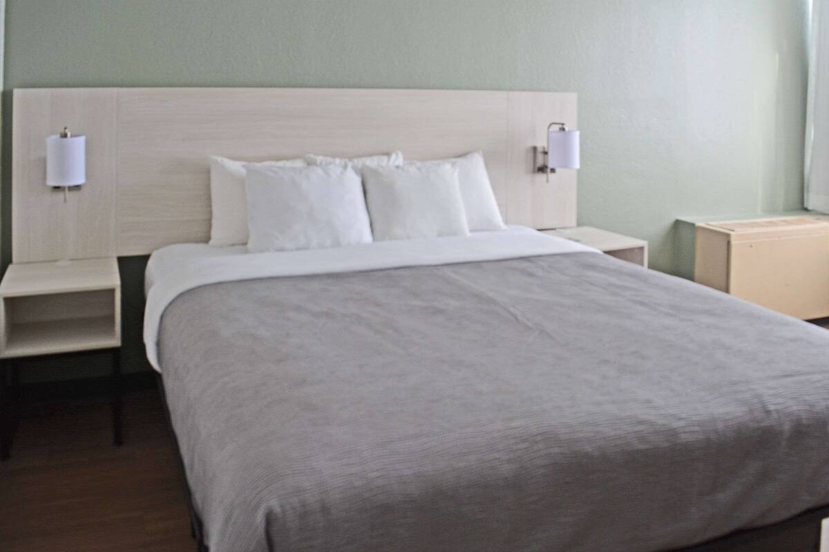 Cama bem arrumada com colcha cinza e almofadas brancas, ladeada por duas mesas de cabeceira com abajures.