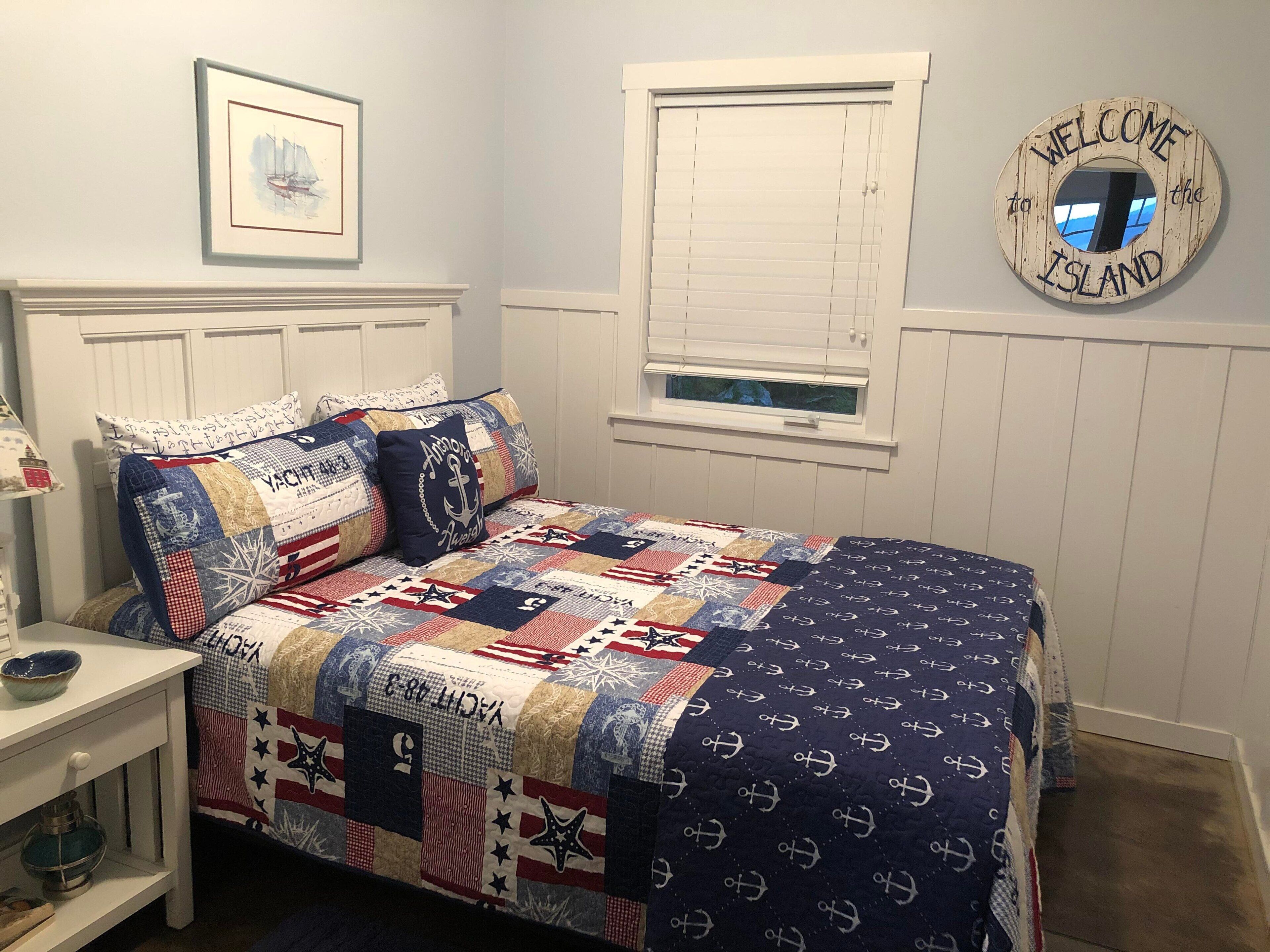 Une chambre à coucher avec une couette bleu marine ornée de motifs d’ancres, une tête de lit blanche, une petite fenêtre avec des stores blancs, un tableau encadré accroché au mur et une pancarte de bienvenue sur le mur.