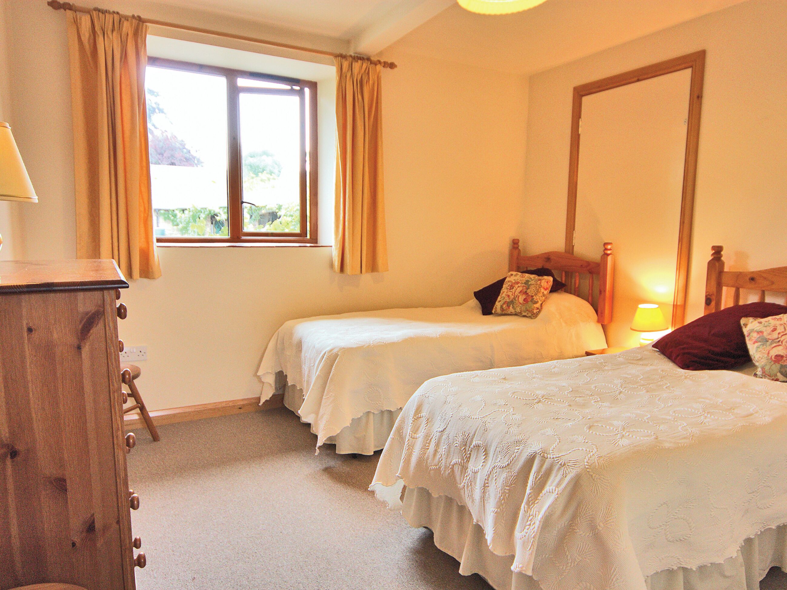 A bedroom with two single beds, a wooden dresser, a window with curtains, and a mirror on the wall.